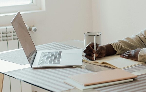 Eine Person schreibt in ein Heft. Vor ihr steht ein Laptop.